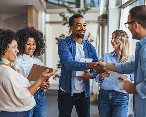 Fünf Personen in einer ungezwungenen Büroumgebung, lächelnd und sich die Hände schüttelnd. Eine Person hält ein Klemmbrett, eine andere ein Tablet.