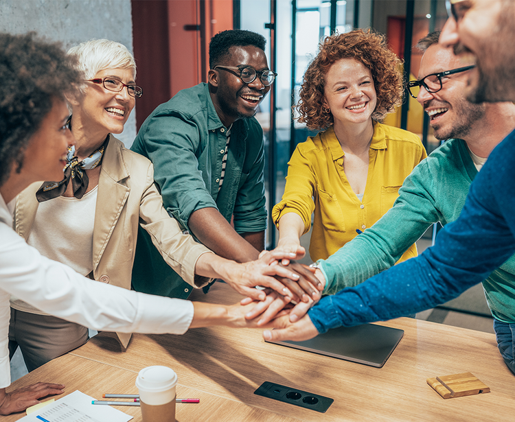 Six people place their hands together over a wooden table in a modern office, symbolizing teamwork and unity. A laptop, coffee cup, and pens sit nearby.