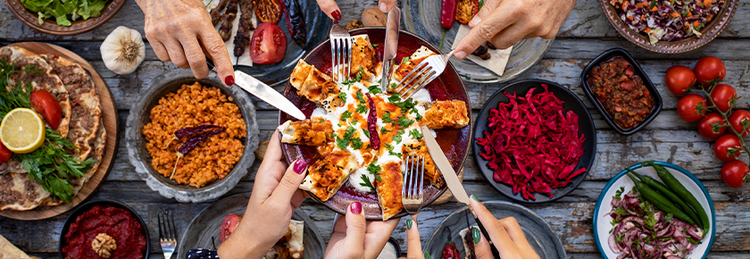 Overhead view of people sharing a communal meal with flatbread, grilled skewers, dips and colourful side dishes, highlighting social dining and shared plate food trends.