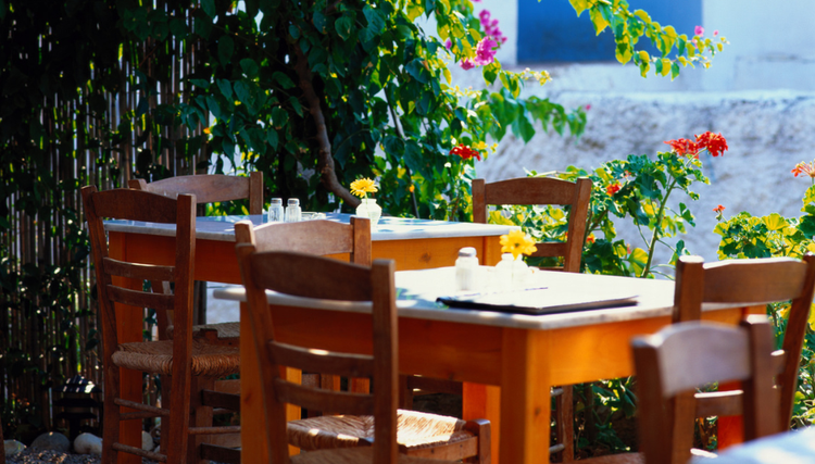 An outdoor dining area with wooden tables set with yellow flowers, surrounded by lush greenery. A cozy and inviting atmosphere.