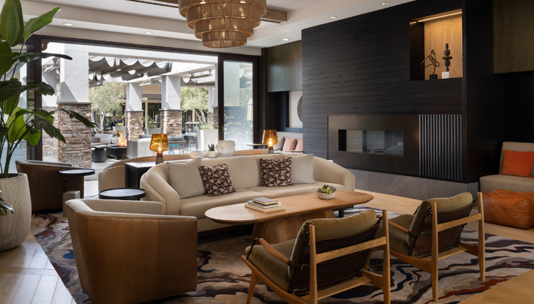 A stylish hotel lobby with warm tones, featuring cozy seating, a black fireplace, and large glass doors opening to an inviting outdoor patio with a fire pit.