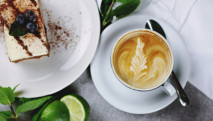 A cup of coffee with leaf-shaped latte art, served with a slice of cake topped with blueberries and mint, alongside lime halves on a white table.