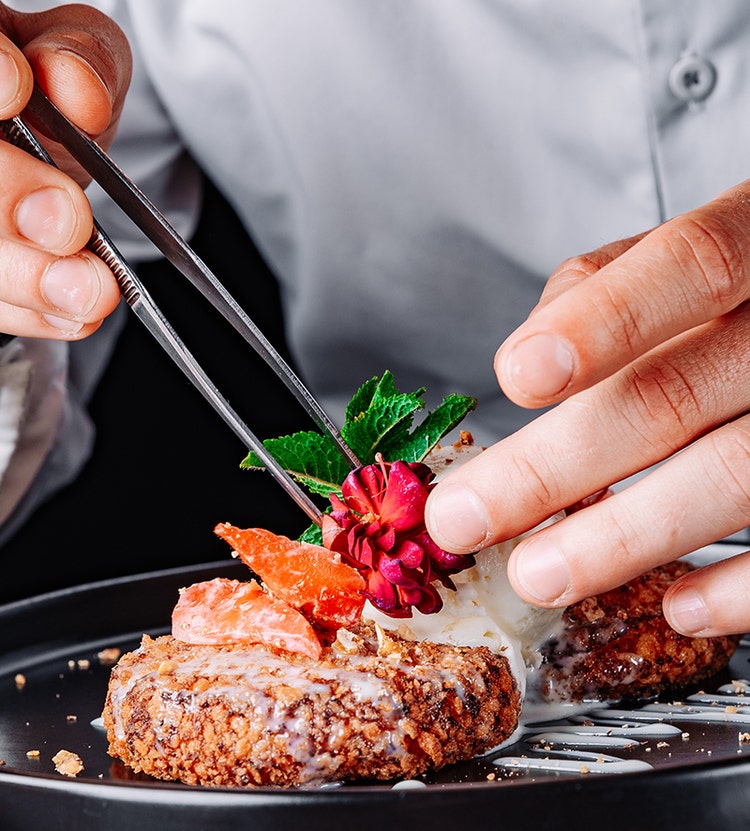 A dessert plate with a crumb-coated treat, drizzled with white sauce, garnished with a red flower, green leaves, and fruit, placed delicately using tweezers.