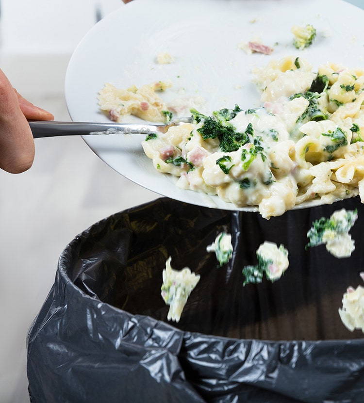 A person scraping leftover food from a white plate into a trash can lined with a black garbage bag.