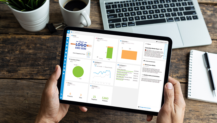 A person holding a tablet with a business dashboard showing colorful charts and graphs, placed above a wooden table with a coffee cup and laptop in the background.