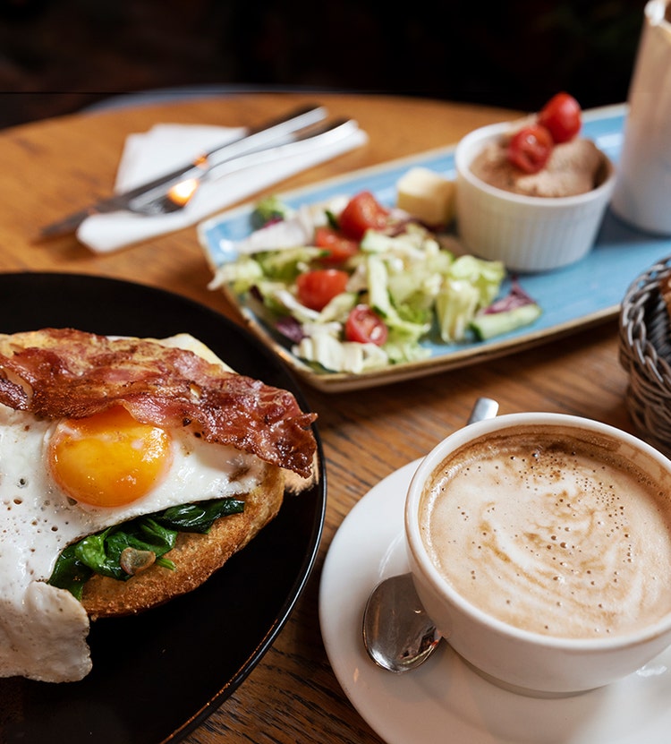 A wooden table with a breakfast spread, featuring a plate of toast topped with a fried egg, crispy bacon, and spinach, alongside a cup of coffee with frothy milk and a small salad in the background.