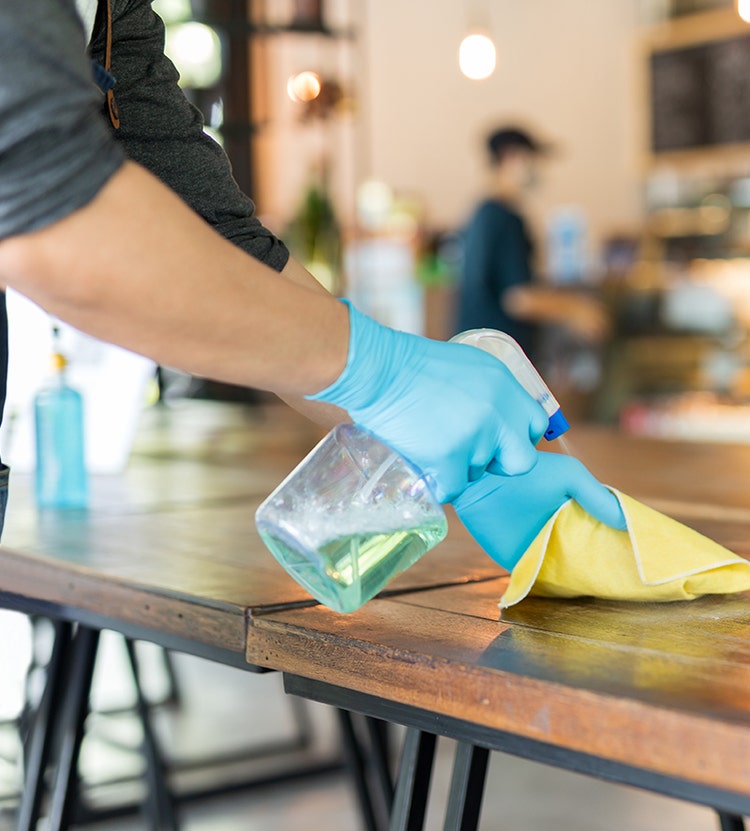 A person wearing blue gloves cleans a wooden table using a green cleaning spray and a yellow cloth in a café setting with a blurred background.