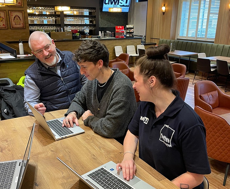 Three people with laptops at a cafe table, surrounded by warm lighting and a bar area in the background.