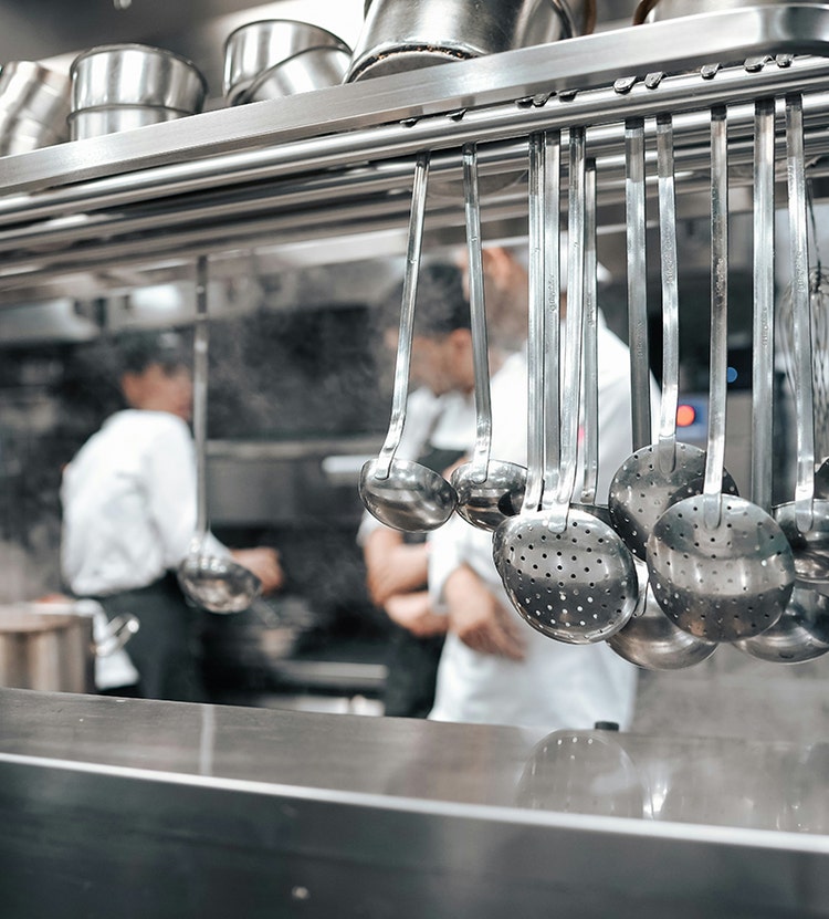 A professional kitchen with stainless steel ladles and spoons hanging on a rack, and two chefs in white uniforms working in the background. Pots and pans are neatly arranged on a shelf above.