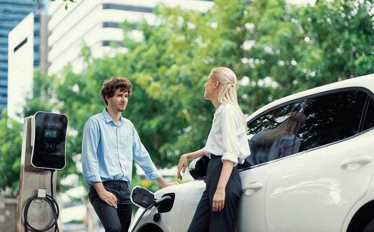 Two people standing next to an electric car charging at a station with a display showing 59%. Trees and buildings can be seen in the background.