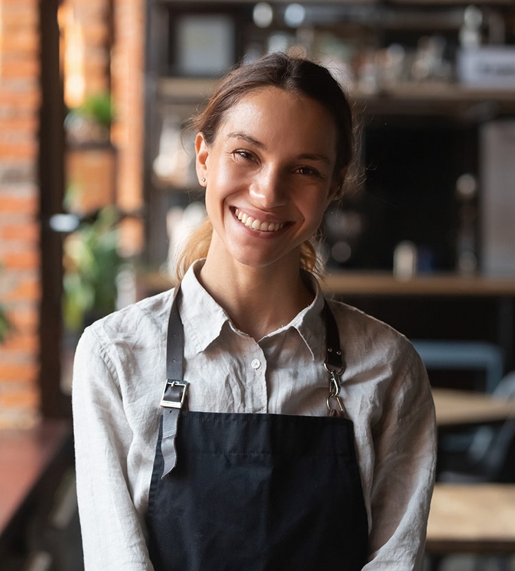A person in a white shirt and black apron standing in a café or restaurant setting with blurred tables, chairs, and shelves in the background.