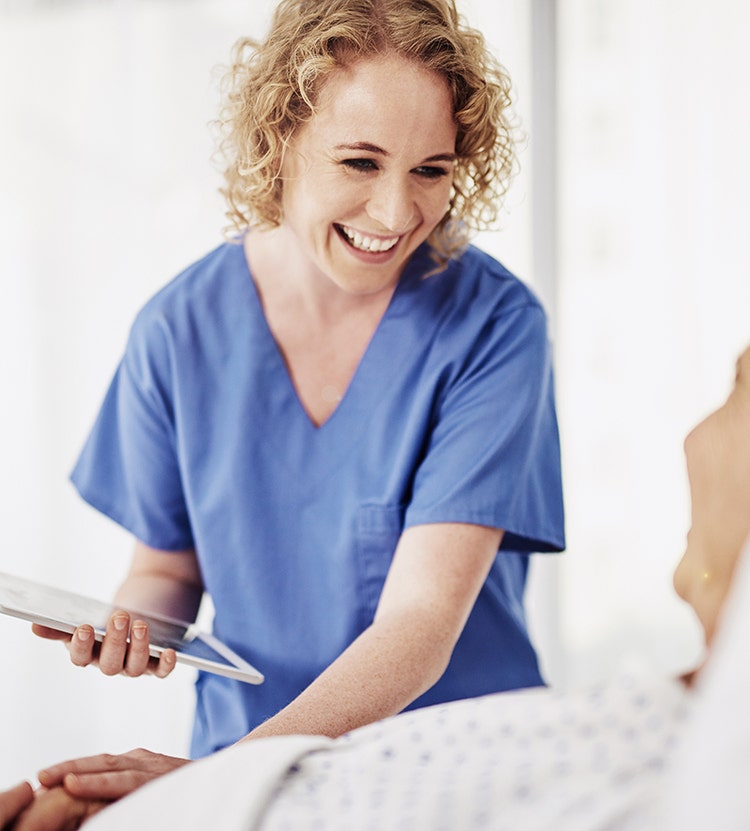 A healthcare professional wearing blue medical scrubs holds a tablet while interacting with a patient lying in a hospital bed.