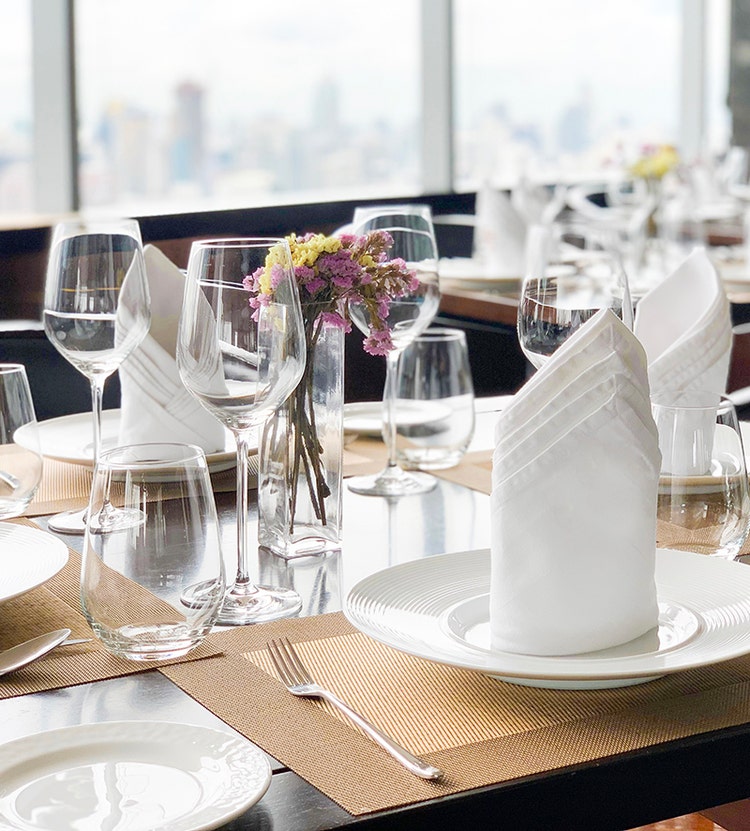 A dining table elegantly set with white plates, folded napkins, wine glasses, and colorful flowers in a small vase, with a view of a cityscape visible through large windows.
