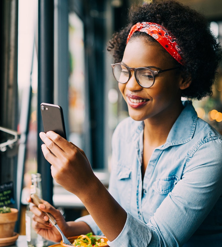 A person wearing a light blue denim shirt with a red headband sitting at a table in a cozy restaurant or cafe, holding a smartphone in one hand and a fork in the other. A plate of food and a bottle of beverage are on the table. The background features large windows and blurred lights.