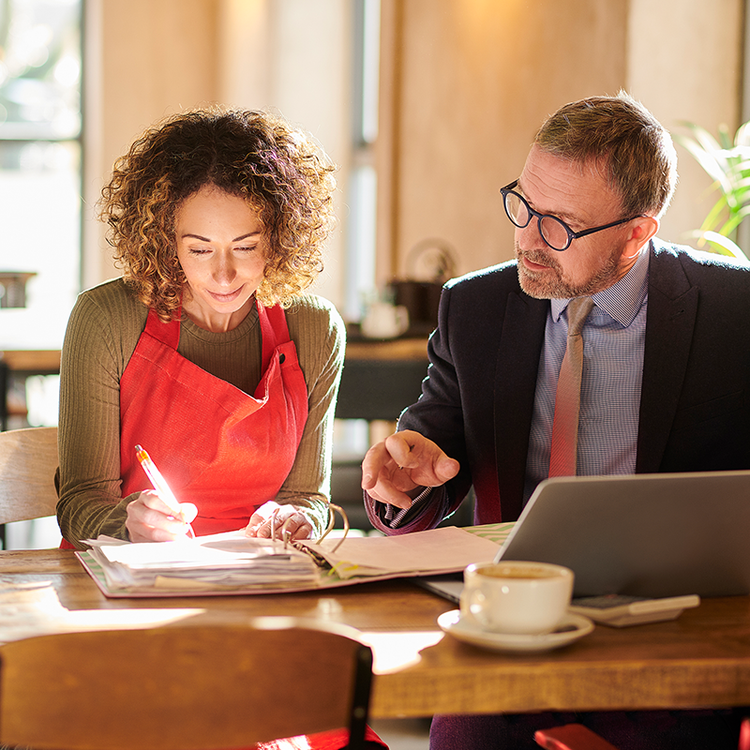 Woman in a red apron reviewing paperwork with a business adviser using a laptop at a café table.