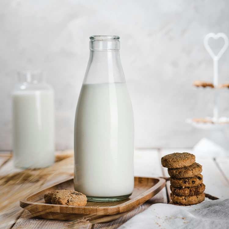 Glass bottle of milk on a wooden tray beside a stack of cookies on a bright table.
