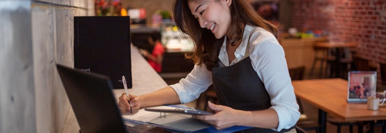 A woman seated with a laptop in front of her, a tablet in one hand, and a pen in her other hand in a cozy atmosphere.