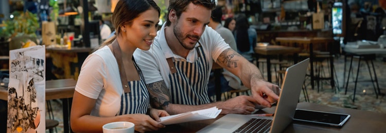 Two people in striped aprons sit at a wooden table in a café, focused on a laptop screen. Papers and a cup are also on the table.