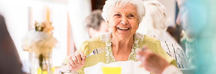 An older woman smiles while seated at a table enjoying her food in a care home