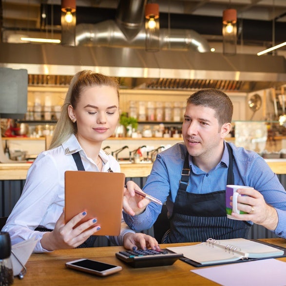 A man and woman are seated at a table, engaged with a tablet in front of them, sharing ideas and collaborating.
