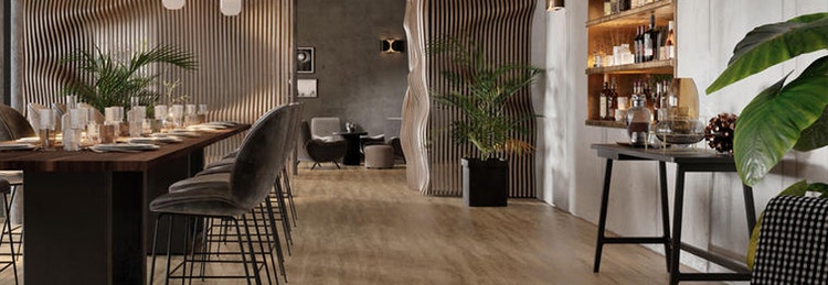 Modern dining area with a long wooden table, high chairs, pendant lights, and a bar shelf. Wooden floors, plants, and textured walls create a cozy atmosphere.