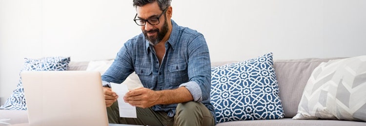 A man seated on a couch, working on a laptop while holding a piece of paper in his hand.