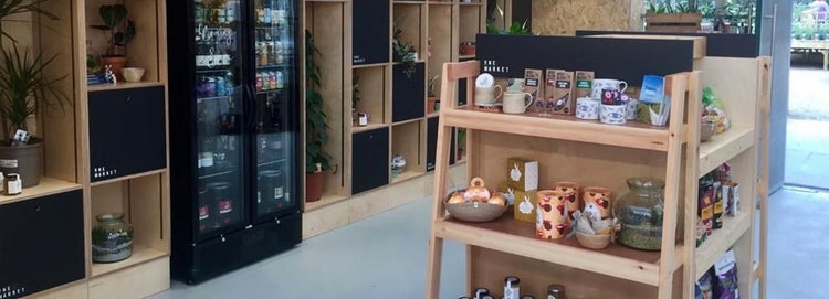 Small shop furnished with wooden shelves selling small gift items, plants, and a fridge of canned drinks