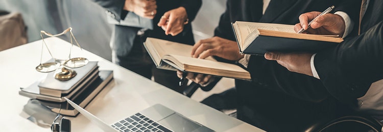 Three people in formal attire discuss open books at a desk with a laptop and scales of justice.