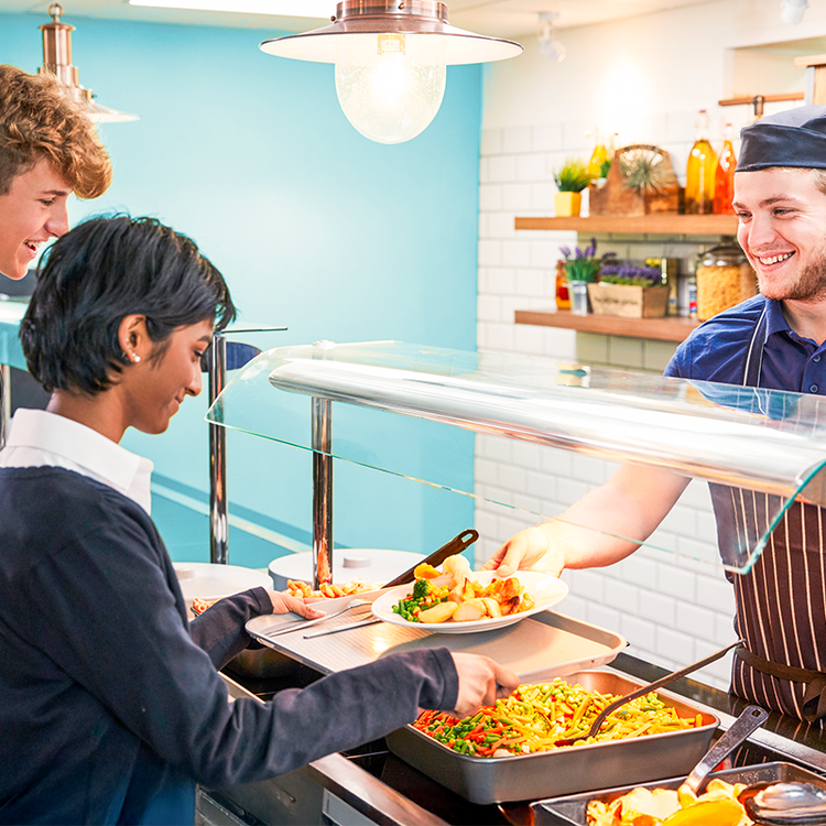 School catering staff serving a hot lunch to students at a cafeteria counter.