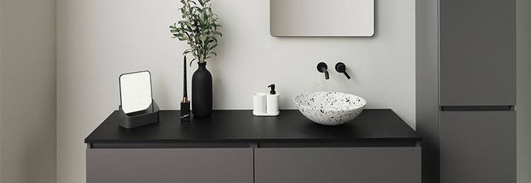 Modern bathroom with a dark gray vanity, black fixtures, a round terrazzo sink, a wall-mounted mirror, and minimalist decor, including a plant in a black vase and a small mirror.