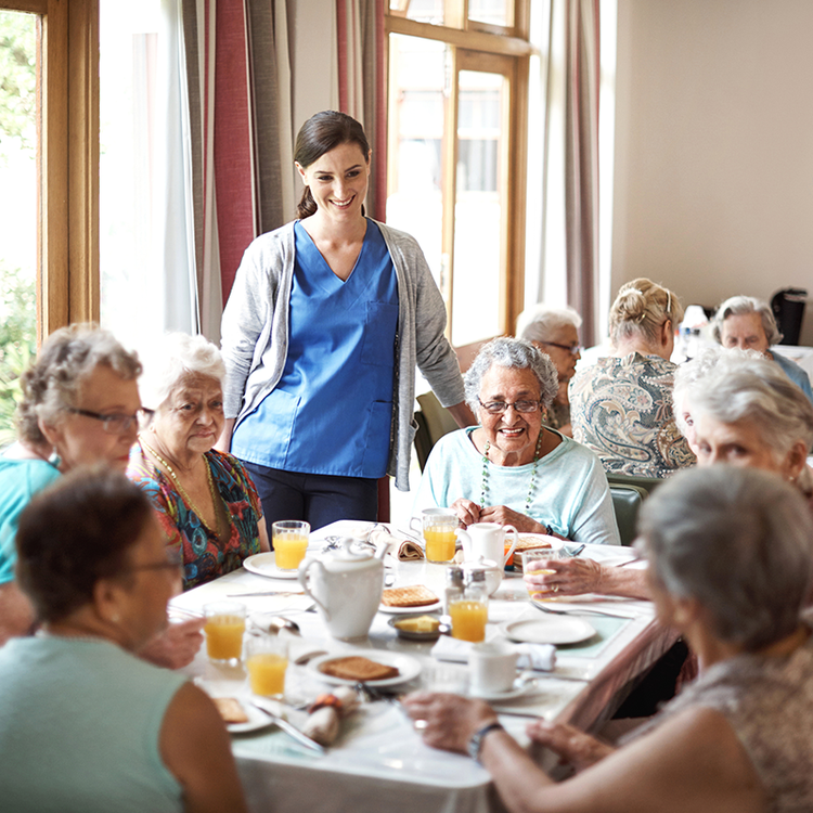 A woman carer serves food to a group of smiling elderly people at a communal dining table