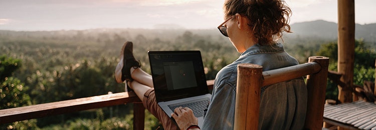 A woman seated on a chair on a patio overlooking a green landscape, focused on her laptop.