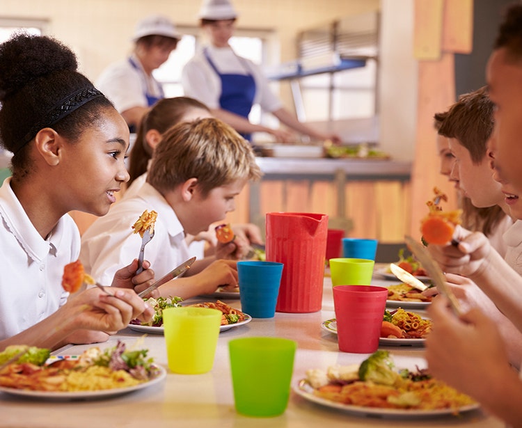 Group of young kids seated at a long table and eatiing together with chefs in the background