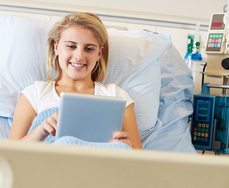 A person lies in a hospital bed under a light blue blanket, holding a tablet, with medical equipment visible in the background.