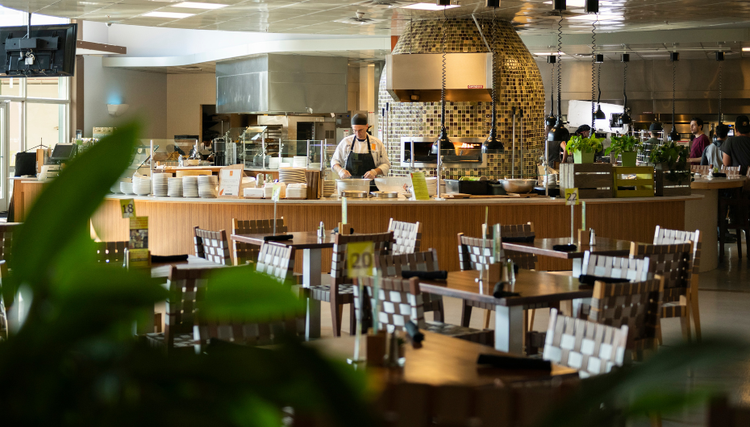 A well-organized restaurant with neatly arranged tables and chairs. In the background, a chef is preparing food in an open kitchen with tiled decor and stacked plates.