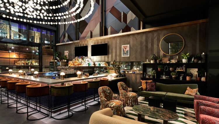 A stylish bar and lounge featuring a large bar counter with stools, modern decor, geometric-patterned walls, and an intricate spherical light fixture.