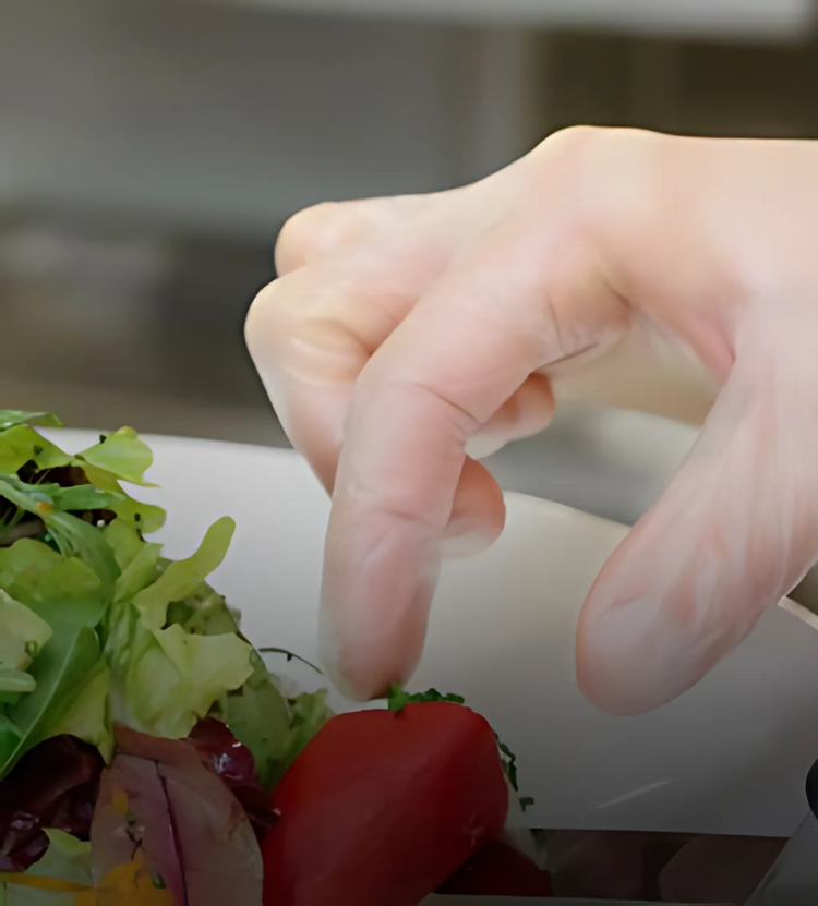 A gloved hand placing a small red tomato on a plate of mixed salad greens, ensuring hygienic food handling