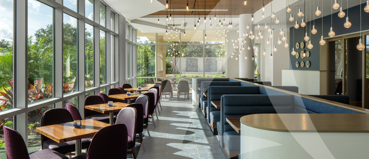 A modern dining area with wooden tables, dark upholstered chairs, blue booth seating, and hanging light fixtures, illuminated by natural light from large windows overlooking lush greenery.