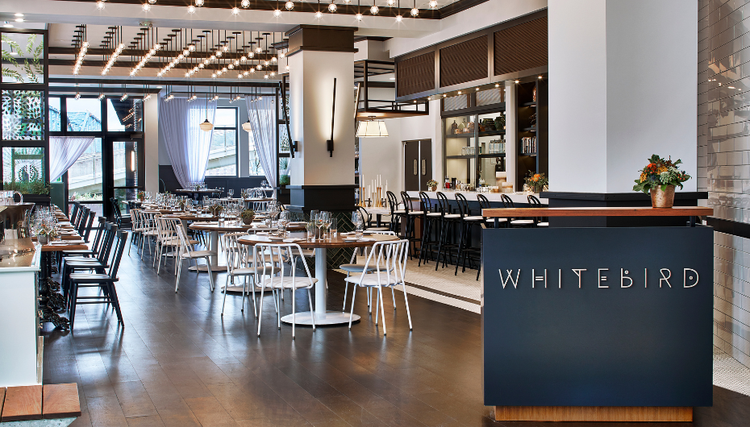 A stylish restaurant named 'WHITEBIRD' with black-and-white chairs, wooden tables, warm lighting, and a bar area in the background.