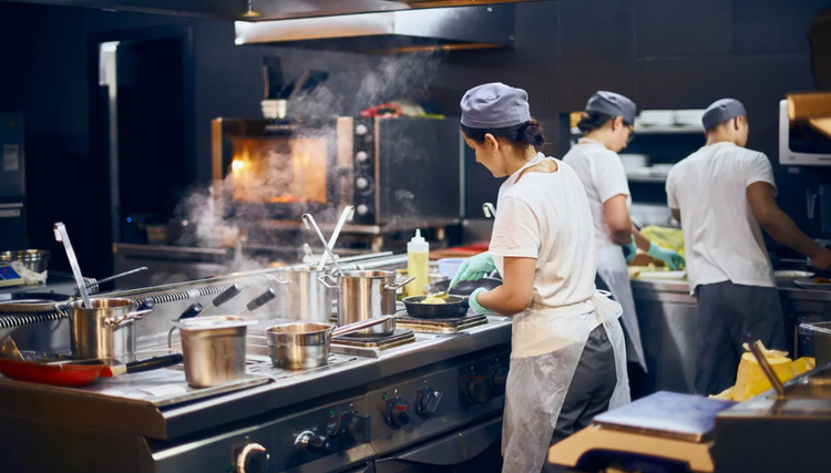 Three chefs working in a kitchen, wearing white shirts, gray aprons, and hats. Steam rises from pots on the stove, and a visible flame glows inside an oven in the background