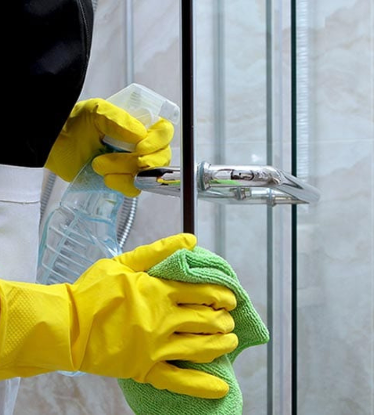 A person wearing yellow rubber gloves cleans a glass door using a spray bottle and a green microfiber cloth.