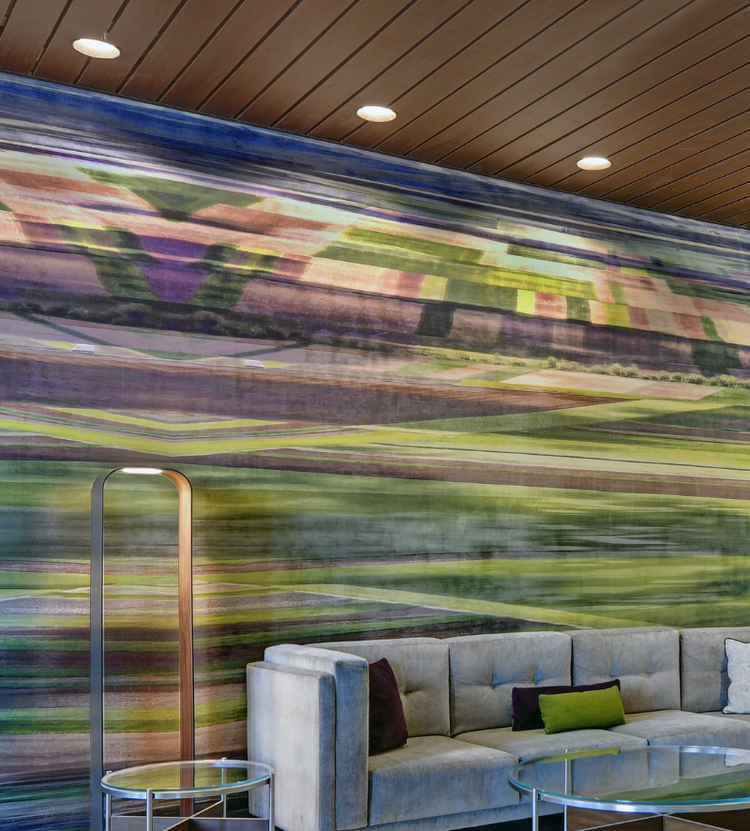 A modern living room with a vibrant abstract mural, a gray sectional sofa, and warm wood-paneled ceilings with recessed lighting.