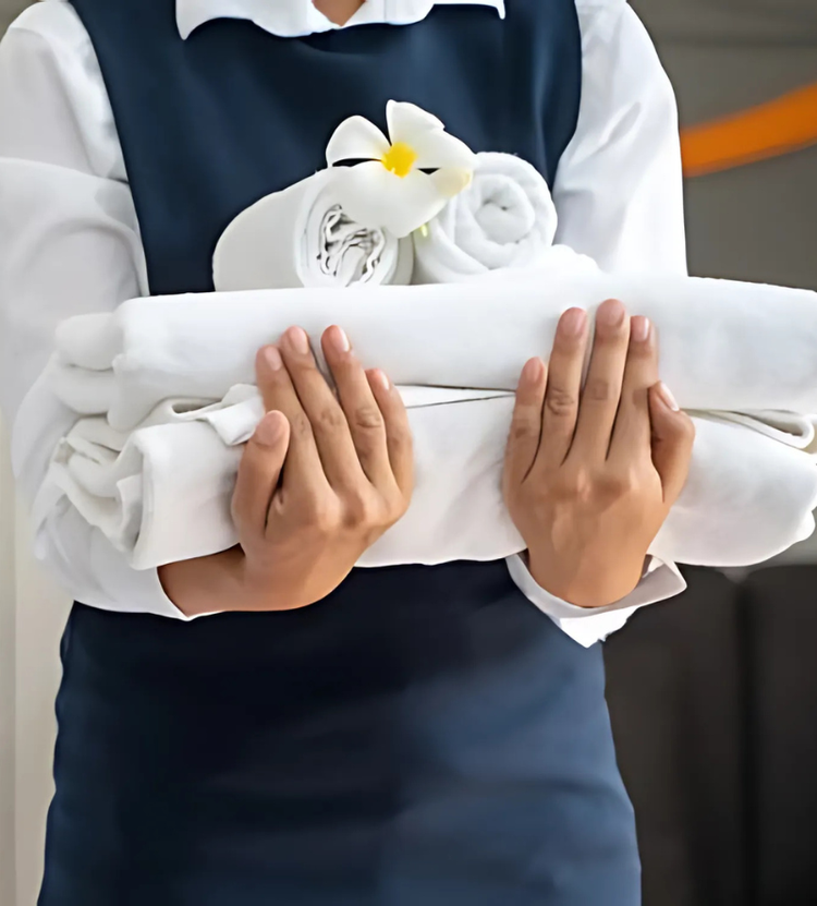 A person in a white shirt and dark blue vest holds a neatly stacked set of white towels. A rolled towel with a white flower rests on top, evoking a sense of cleanliness and hospitality.
