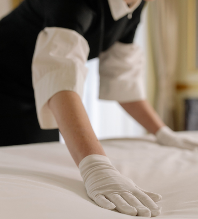 A person in a black and white uniform with white gloves smooths a bed sheet, highlighting attention to detail in housekeeping.