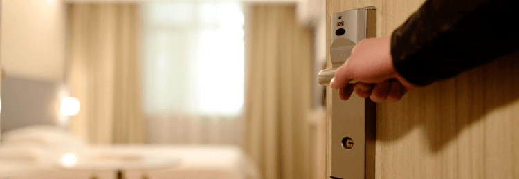 Hand opening the door to a hotel room to show a small table, bed, and window