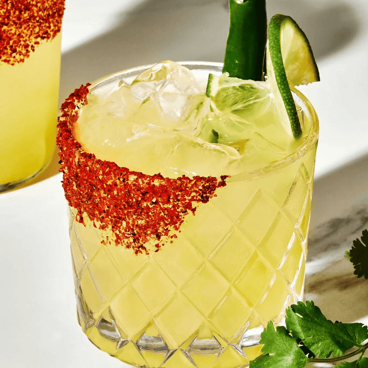 A refreshing yellow cocktail in a textured glass with a chili salt rim, garnished with a green chili pepper and a lime wedge, placed on a white marble surface with fresh cilantro leaves nearby.