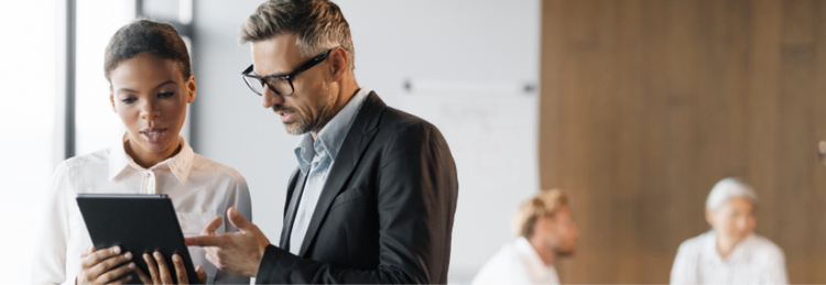 Two people stand and discuss while looking at a tablet in a modern office. In the background, two others sit and talk.