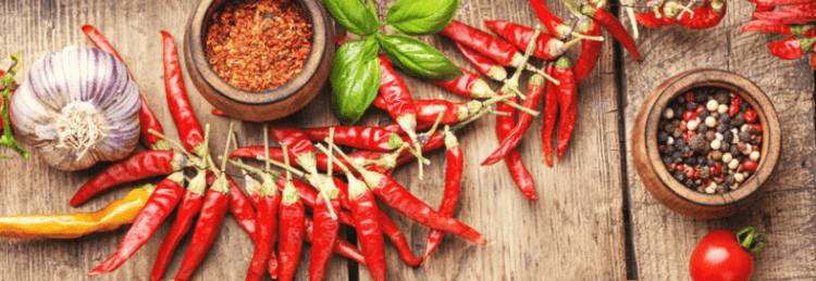 A rustic wooden table filled with fresh red chili peppers, dried spices, garlic, cherry tomatoes, and a wooden bowl of spicy red sauce, accompanied by a wooden spoon.