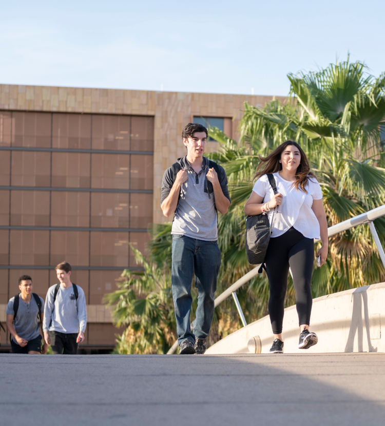 Four individuals walking along a paved pathway, carrying backpacks in a campus with a modern building and palm trees in the background.