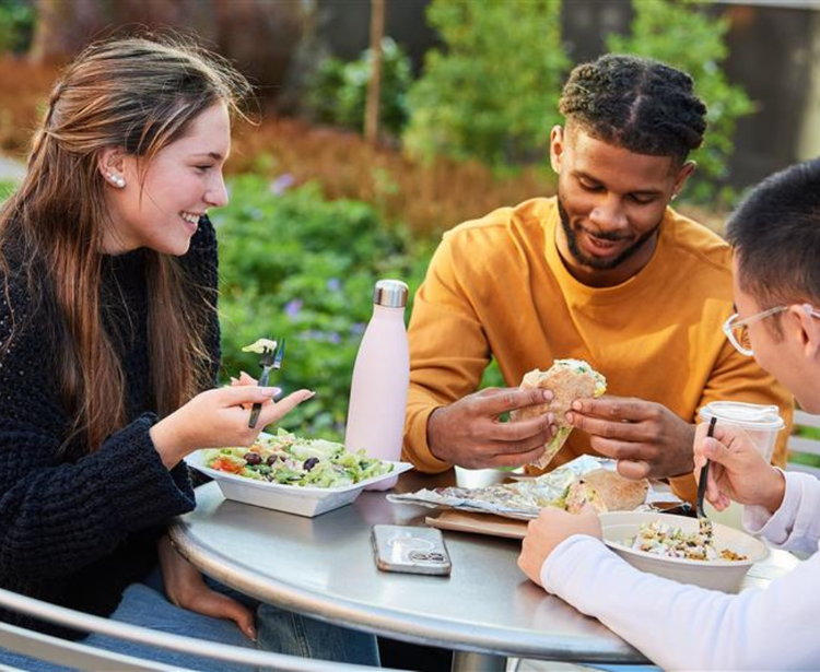 Three people dining outdoors at a table, enjoying a meal together with a pink water bottle and a smartphone nearby.